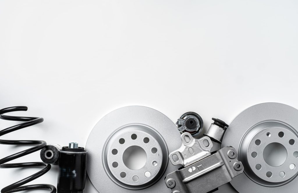 A flat lay of new metal auto parts, including brake discs, suspension springs, and other components, arranged on a clean grey background.